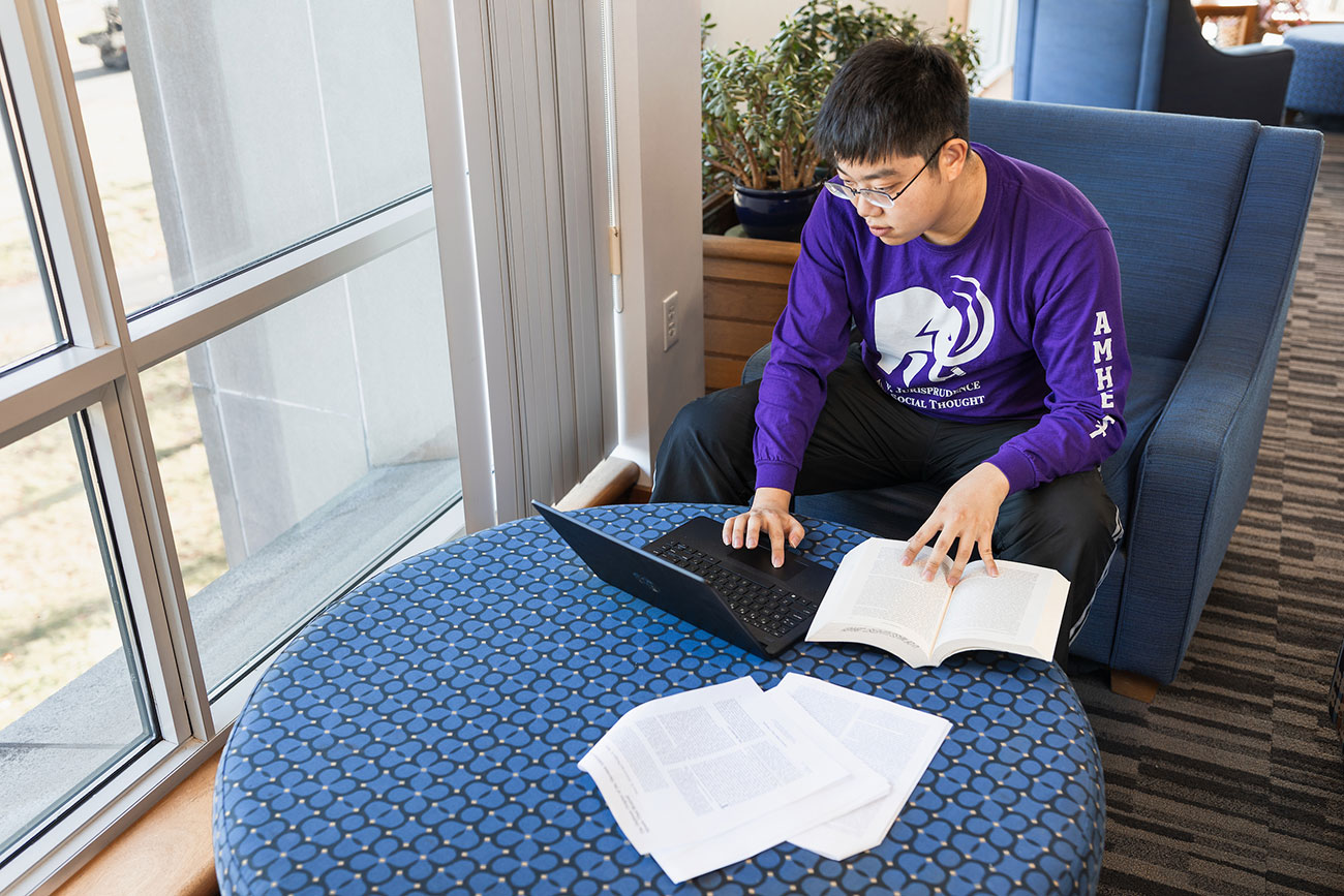A young man in a purple tshirt with a lap top studies at a desk.