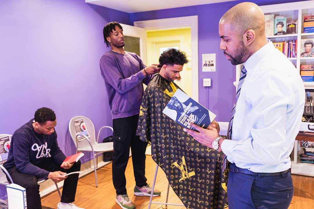 Prof. Roberts reads from a book while one student gives another a haircut as a second student waits for his turn