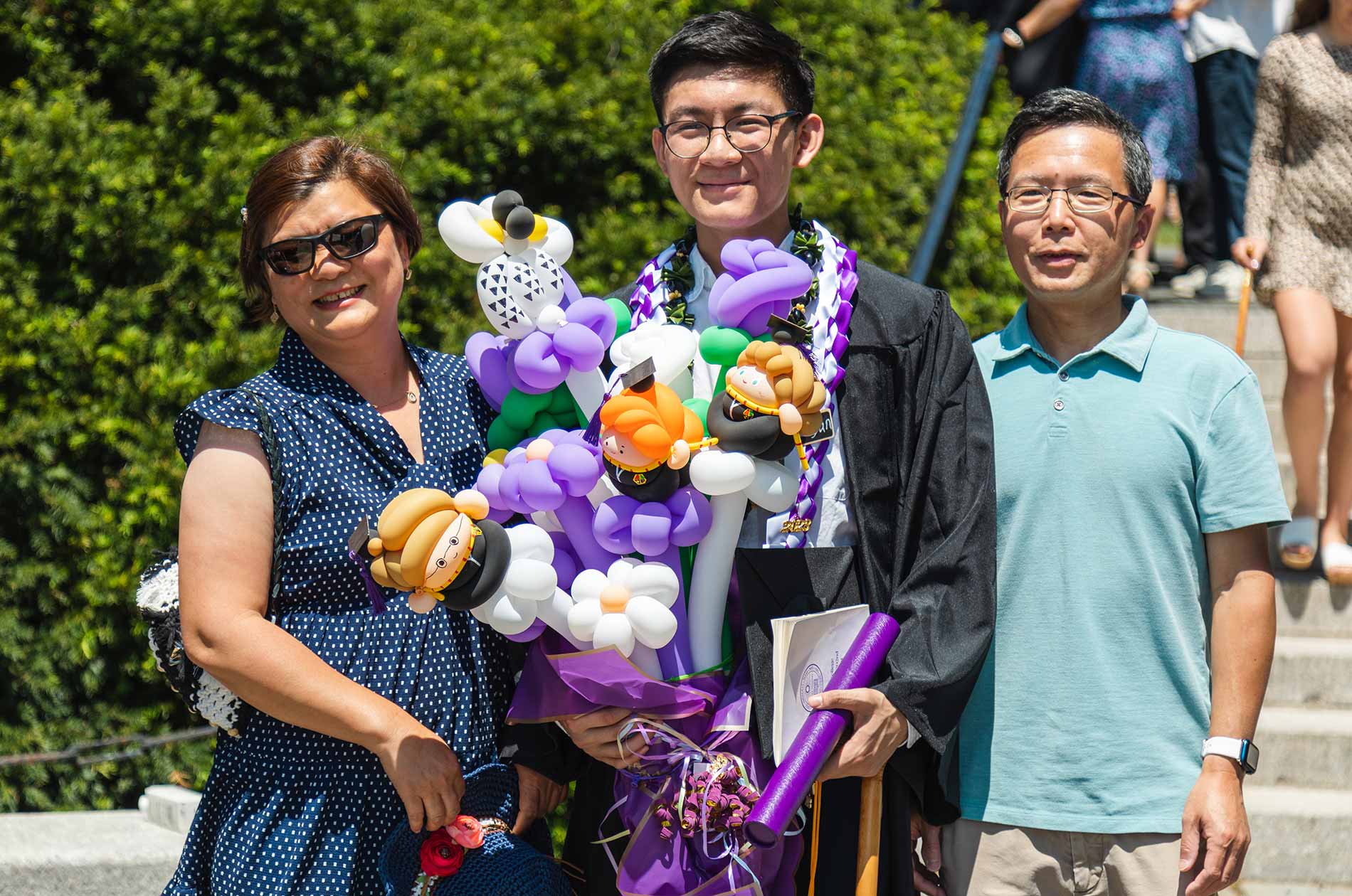A graduate, holding a large bouquet of balloons. poses with family members