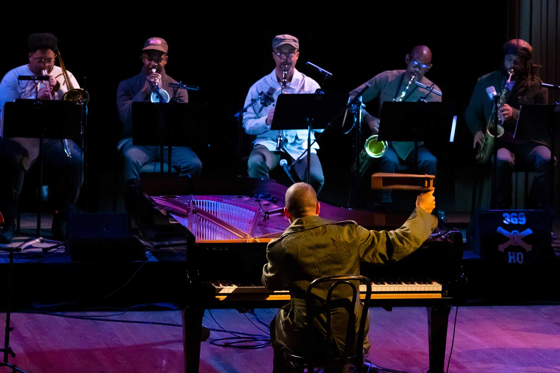 A man plays a piano facing a line of men playing wind instruments.